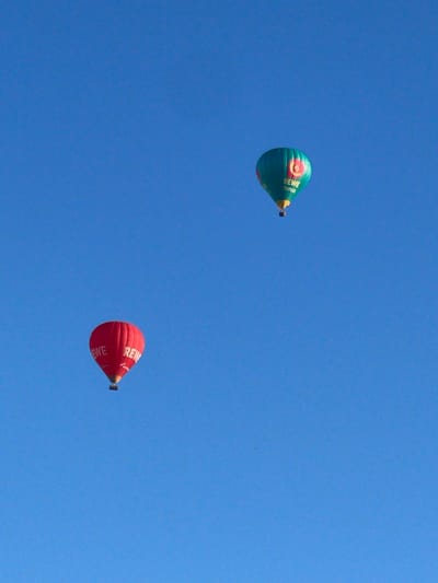 Two hot air balloons are visible against a clear blue sky. One balloon is red with "EWE" written on it in white letters. The other balloon is teal with "REWE Bonus" written in yellow letters. Both balloons appear to be in flight. The image does not provide any information about the city of Hamburg, Germany or the location within it.