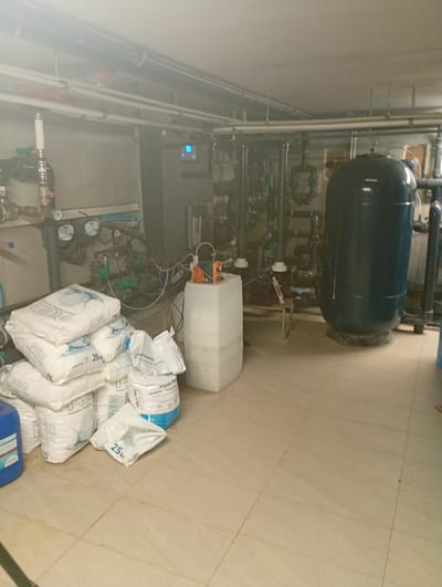 The image depicts an indoor mechanical room, likely for a pool or water treatment facility, located in Nowa Wieś Szlachecka, Poland. The room is filled with various equipment including pipes, a large dark blue cylindrical tank, a grey electrical control panel with a blue display, and several large white bags of chemical products, some labeled "25kg" and "AQUA PRO". A tall white plastic container is also visible, with some orange tubing connected to it. The floor is tiled in a light beige color. There are no people present in the scene. The environment appears to be dimly lit with artificial lighting, and there is some dust or fog in the air, particularly in the upper portion of the image.