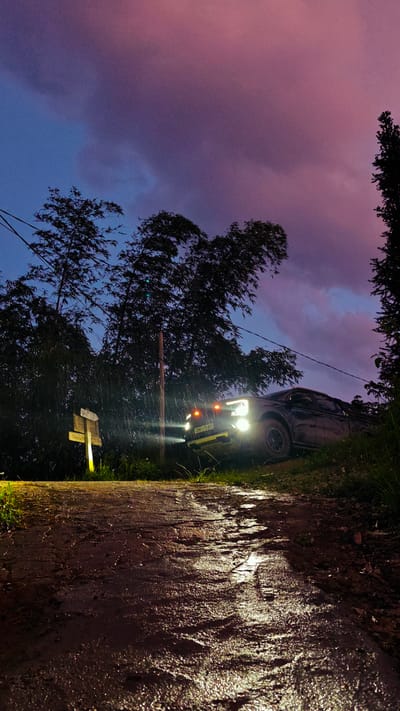 The image depicts a rural scene in Hoàng Su Phì Commune, Vietnam, at twilight or night. A dark-colored pickup truck with illuminated headlights is parked on a muddy dirt road, facing uphill. The road surface is reflective, suggesting recent rain. A wooden signpost with "Văn Ly" on it is visible to the left of the truck, also illuminated. Tall trees line both sides of the road, silhouetted against a sky with a gradient of colors from blue to pink and purple. Rain is falling, visible as streaks of light near the truck's headlights. The license plate of the truck shows "29C-175.81."