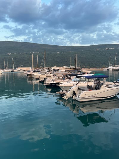 A vertical shot captures a serene marina scene in Đenovići, Montenegro, with a line of boats docked along the water. The boats, varying in size and style, are mostly white with dark trim, and many are equipped with outboard motors. One prominent boat in the foreground has "Cap Camarat" written on its side, and another visible engine bears the "Honda" logo. A sign on one of the boats advertises "RENT A BOAT" with a phone number and website, indicating a commercial operation.
The water is calm and reflects the sky, the boats, and the lush green mountains that rise steeply behind the marina. The mountains are densely covered in trees, and small houses are scattered along their slopes, hinting at a developed but still natural landscape. The sky is partly cloudy, with a mix of bright white cumulus clouds against a pale blue background, suggesting a pleasant, possibly late afternoon, time of day. The overall impression is one of tranquility and a typical summer day at a coastal resort. No people are clearly visible in the image, and the focus is on the boats and the surrounding environment.