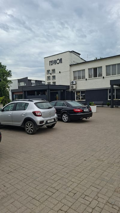 The image shows a street scene in Smaliavichy, Belarus, featuring a building with the Cyrillic word "ГЕРМОН" visible on its facade. Two cars are parked in front of the building: a silver Renault Sandero Stepway and a black Mercedes-Benz E-Class. The building appears to be a commercial establishment, with large windows and outdoor seating areas. The ground is paved with interlocking bricks, and the sky is overcast.