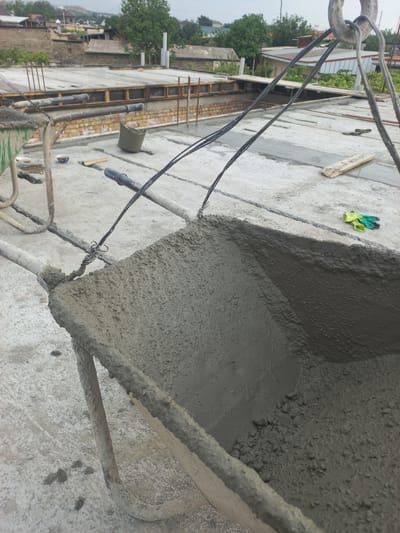 The image depicts a construction site, likely in the city of Khujand, Tajikistan. In the foreground, a wheelbarrow filled with wet concrete is prominently featured, its contents close to overflowing. The wheelbarrow is suspended by thick ropes connected to a heavy-duty hook, suggesting it is being lifted or moved.
In the mid-ground, a concrete surface is visible, with sections of rebar and formwork indicating ongoing construction. A bucket, also filled with concrete, rests on the surface. Further in the background, buildings with tiled roofs and trees are visible, suggesting a semi-urban or suburban setting. A pair of yellow and green work gloves are placed on the concrete surface.
The overall scene is one of active construction work, with a focus on the transportation and pouring of concrete. The environment appears to be an outdoor setting under an overcast sky.