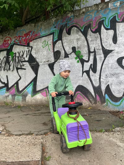 A young child wearing a green jumpsuit and a patterned beanie is playing with a green and purple toy car on a cracked asphalt surface. The child is holding onto the steering wheel of the toy car. Behind the child and toy car is a concrete wall covered in vibrant graffiti. Overhanging trees with green leaves are visible at the top of the image, suggesting an outdoor, urban setting. The scene appears to be in Izhevsk, Russia.
