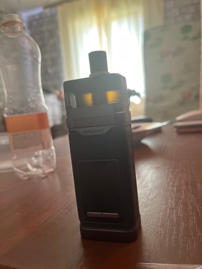 A black vape device with "SMOANT" branding sits on a wooden table. To the left of the vape is a clear plastic bottle with a white label containing orange text and several small air bubbles inside. In the background, a window is covered by light yellow curtains, and a patterned chair is visible to the right. The scene appears to be indoors, potentially in a residential setting in Baykalsk, Russia.