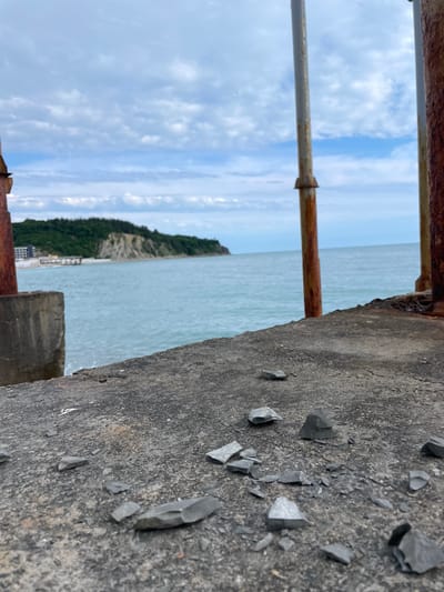 The image depicts a close-up view of a concrete ledge or wall with scattered dark, angular fragments. Rusted metal railings are present in the foreground. Beyond the ledge, the scene opens to a body of water, possibly the sea, with land visible in the distance. The sky is cloudy with patches of blue.

While the image's location is stated to be Olginka, Russia, the provided visual information alone does not confirm this location. Additional contextual clues would be needed to verify this claim.