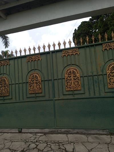 The scene is captured in what is stated as the city of Vieng Phouka, Laos. The primary subject is a large, dark green metal gate, likely a sliding entry gate, viewed from an exterior perspective. The top edge of the gate features a series of golden, pointed finials and decorative horizontal friezes. Below these, the upper section of the gate incorporates three visible arched, gold-colored ornate panels with intricate scrollwork. The lower section of the gate displays a diamond plate texture. A black wheel is visible at the base of the gate, resting on a cracked concrete ground surface exhibiting multiple fractures. Overhead, a light-colored concrete beam or structure spans horizontally across the upper frame. In the background, a cloudy sky is visible, with dense green tree foliage on the right and a palm frond on the left. A thin dark wire crosses the upper right portion of the sky. No individuals are present, and no active interactions are depicted.