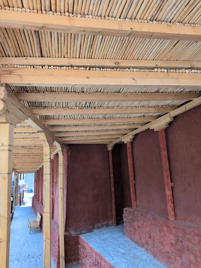 The image shows the interior of a covered walkway with a rustic wooden ceiling constructed of numerous small, parallel wooden poles supported by larger wooden beams. The walls are a reddish-brown plaster, and the floor is paved with light blue stones. In the foreground, a wooden beam is supported by a carved wooden pillar. To the left, the walkway extends into a well-lit area with a paved path and a wooden bench. A person is partially visible at the far end of the walkway on the left. The architecture suggests a traditional or historical building. The image does not provide specific information about the city of Choma, India.