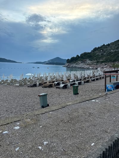 The image depicts a tranquil beach scene in Kamenovo, Montenegro. The foreground is dominated by a textured, grey concrete walkway and a sandy beach lined with rows of empty white beach chairs and umbrellas. A green garbage bin with wheels sits on the sand.
Beyond the beach, the calm, blue sea stretches towards distant, tree-covered mountains. The sky is partly cloudy, with soft sunlight breaking through, suggesting it's either early morning or late afternoon. Several small boats are visible on the water, adding a touch of activity to the otherwise still setting. The overall atmosphere is peaceful and serene, with no people visible on the beach or in the water.
In the background, part of a building with an ice cream freezer is visible, hinting at a nearby establishment. The rock face of the mountain rising behind the beach is covered in lush green vegetation, creating a striking contrast with the sea and sky. The scene evokes a sense of quiet relaxation and natural beauty.