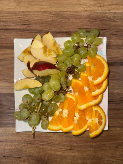 On a wooden surface is a white square plate containing a fruit assortment. The fruit includes green grapes, orange slices, and apple wedges. The scene is likely indoors. The location in Esenyurt, Turkey is not directly discernible from the image.
