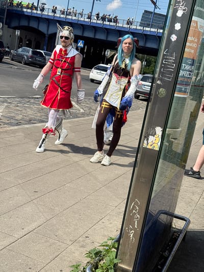 Two individuals in elaborate costumes are walking on a paved sidewalk. The person on the left, dressed in a red and gold outfit resembling a character from a video game, is wearing white gloves and knee-high white boots with red bows. They have on sunglasses and a wig. To the right, a person with blue hair and red horns is also in costume, wearing a dark brown and white outfit. They are walking in the same direction. Behind them, a blue bridge spans over a street with cars. The scene appears to be in a public urban area, possibly during an event or convention, given the costumes. A glass structure, likely a bus stop or information kiosk, with graffiti and posters, is to the right of the frame. Based on the visible signage and architectural style, the location is identified as Poznań, Poland.