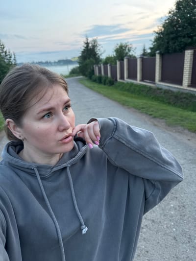 A young woman with light brown hair pulled back stands on a paved road, facing left and looking away from the camera. She is wearing a grey hooded sweatshirt and has her right hand up to her mouth, with her fingers touching her lips. Her fingernails are painted pink. The road appears to be in a rural or suburban setting, with trees and a fence visible in the background. The sky is overcast and appears to be either dawn or dusk. The location is identified as Gorniy, Russia.