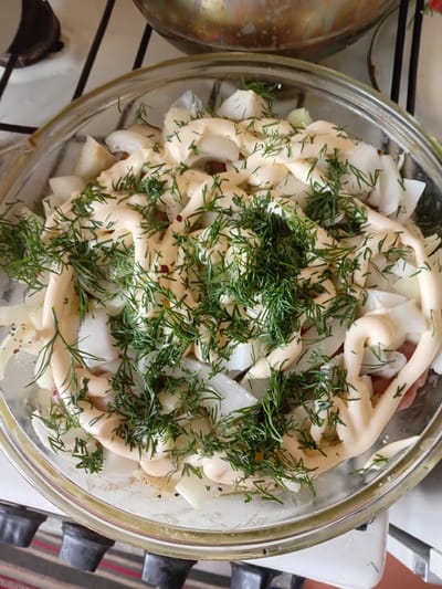 A close-up, overhead shot shows a glass bowl filled with a salad. The salad appears to consist of chopped white vegetables, possibly onions or potatoes, interspersed with pieces of meat, perhaps chicken or pork, which are a light pinkish-brown color. The dish is generously dressed with a creamy white sauce, likely mayonnaise, which has been drizzled in wavy lines across the ingredients. Fresh dill is scattered liberally over the top, adding a vibrant green contrast to the lighter colors of the salad and dressing. Small flecks of red, possibly chili flakes or paprika, are also visible. The bowl sits on a stovetop, indicated by the black grate and white enamel surface. To the left of the bowl, a portion of a shiny metal pot is visible. The overall impression is of a homemade dish being prepared. The lighting suggests it might be daytime, but no specific time of day can be determined. The setting appears to be a kitchen. There is no visible text in the image.