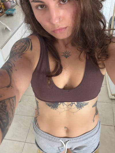 A selfie of a woman indoors. She has brown hair and brown eyes. She is wearing a brown ribbed sports bra and gray sweatpants. She has multiple tattoos on her torso and arm. The background appears to be a tiled floor and a partial view of a room. There is no evidence to suggest that this image was taken in Kiryat Bialik, Israel.
