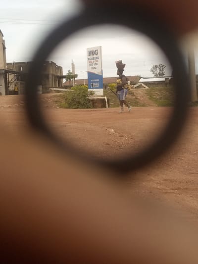 The image captures an outdoor scene in Kwang, Nigeria, viewed through a circular, blurred lens, likely from a camera or optical device. In the foreground, obscuring a large portion of the frame, is a dark, blurred ring. The background reveals a street scene.

The focal point of the image is a person walking, carrying items on their head and holding a child or another item against their body. The person is wearing blue shorts and a dark top. They are walking on a dirt road, which appears to be uneven with small stones and patches of dry earth.

To the left of the person is a tall, white sign with text that is partially visible and difficult to read clearly due to the overall blur. It appears to be an advertisement or informational sign, possibly for a company or service. Behind the sign and the person, there are low-rise buildings and vegetation. The sky is overcast, suggesting a cloudy day. The environment appears to be a rural or semi-urban setting within Kwang, Nigeria.