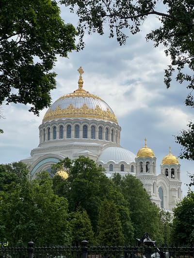 The image captures a view of the Saint Nicholas Naval Cathedral in Kronstadt, Russia, framed by lush green trees and a cloudy sky. The cathedral is a prominent white structure with multiple domes. The main dome is large and elaborately decorated with gold trim, topped by a golden cross. Smaller golden domes, also topped with crosses, are visible on attached bell towers. The architecture features arched windows and detailed white stonework. In the foreground, a black decorative fence is visible, suggesting a public space or park. The surrounding trees are full of green leaves, indicating it is likely summer. The overall scene is tranquil and majestic, highlighting the grandeur of the cathedral against a natural backdrop.