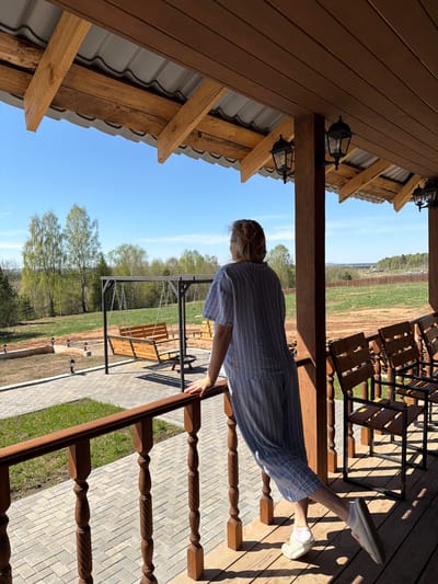 A woman in a blue and white striped dress stands on a wooden porch, leaning on the railing. She faces a yard with a swing and landscaping in what appears to be a rural or suburban setting. The location may be Kamennoye, Russia, based on the presence of wooden buildings and surrounding greenery. Overhead is the roof of a wooden structure, possibly a house or cabin, with exposed beams and a gray metal roof. Several wooden chairs are visible further down the porch. The sky is blue and sunny.