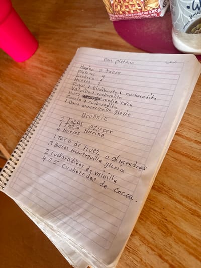 The image shows a handwritten recipe in Spanish, likely for banana bread ("Pan platano") and brownies ("Bronnie"). The recipe ingredients are written on a lined notebook paper. The notebook is lying on a wooden surface next to other objects, which are out of focus.