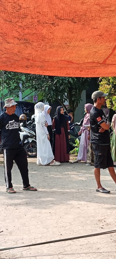 The image was taken in Cangkuang, Indonesia at the coordinates -7.065, 107.552.  There are seven people standing in a group on a paved ground. Six of the people are wearing traditional Indonesian clothing. Two women are wearing long white dresses with headscarves. One woman is wearing a long maroon dress with a black headscarf. Another woman is wearing a long purple dress with a headscarf. Two other women are obscured from view. One man is wearing a black shirt with the words "Durban" and a black baseball cap. Another man is wearing a black t-shirt with the word "TRUE" on the front and a black baseball cap.  A motorbike is parked behind them. There is a large orange tarpaulin above the group, and a tree is to the left.  The ground is covered in leaves and twigs.  One man is looking at the camera.  Another man is looking off to the left. The rest of the people are looking down. The men are standing with their hands at their sides or in their pockets. The women are standing with their hands crossed in front of them. The men and women do not appear to be interacting.