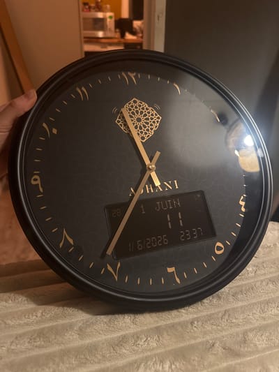A black circular wall clock with golden Arabic numerals and hands is prominently displayed, held by a person's hand in the bottom left corner. The clock face features a subtle black geometric pattern and a distinctive golden Islamic-style geometric design at the 9 o'clock position. The brand name "KHANI" is vertically inscribed in gold near the center of the clock.

An integrated digital display screen on the clock face shows several pieces of information. The date is clearly visible as "1 JUIN" (June 1) and "1/6/2026", suggesting the image was taken on June 1st, 2026. The digital time reads "23 37", indicating 11:37 PM. However, the analog hands of the clock point to approximately 9:48, showing a discrepancy with the digital time. Additional smaller numbers, "28" and "11", are visible below the main digital display, possibly related to prayer times or other clock functions.

The background on the right consists of a light-colored, textured fabric, likely a blanket or towel. To the left, a blurry indoor setting, possibly a room in a home in Wattrelos, France, is visible with warm lighting and indistinct furniture. The scene captures the clock in detail, highlighting its design and functional elements.