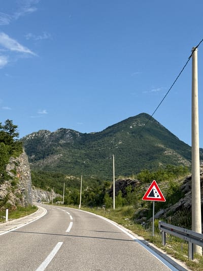 A winding asphalt road curves through a mountainous landscape under a clear blue sky. The road is marked with white dashed and solid lines. On the right side of the road, a concrete utility pole stands tall, with a black wire running diagonally across the sky. Beside the pole is a triangular warning sign with a red border and a black graphic depicting falling rocks, indicating a potential hazard. A metal guardrail runs along the edge of the road. To the left, a steep, rocky cliff face is visible, covered in sparse vegetation. Further back, lush green trees and a large, prominent mountain dominate the background. Several smaller utility poles are spaced along the side of the road. A dark-colored vehicle is visible in the distance, approaching on the road. The overall scene suggests a remote, mountainous area, likely in a region prone to rockfalls, such as the area around Dragalj, Montenegro.
