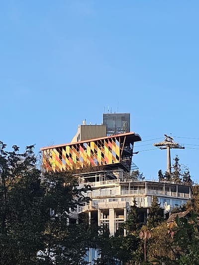 This media file captures a modern cable car station situated on a hillside under a clear blue sky in ქვედა სამება, Georgia. The main subject is a building that appears to be part of the cable car infrastructure, possibly a station or an observation deck. Its most striking feature is a vibrant, multi-colored facade on its sloped side, composed of geometric patterns, specifically diamond or square shapes in hues of red, yellow, blue, black, white, and orange, creating a stained-glass-like effect.
Above and to the left of this colorful structure, a cable car pylon stands against the sky, with cables extending from it, indicating the presence of a cable car line. No individual cable car cabins are visible.
In the background, nestled into the hillside, are several contemporary multi-story buildings, likely residential or commercial, featuring balconies and numerous windows. Lush green trees and dense foliage are visible in the foreground and interspersed among the buildings, highlighting the natural, possibly mountainous or hilly, landscape.
The time of day is clearly daylight, likely late morning or early afternoon, given the bright, even lighting and the absence of long shadows. The weather is sunny with an unclouded, clear blue sky. There are no people visible in the scene, and thus no specific activities are explicitly depicted beyond the implied function of the cable car system for transportation. No visible text is present in the image.