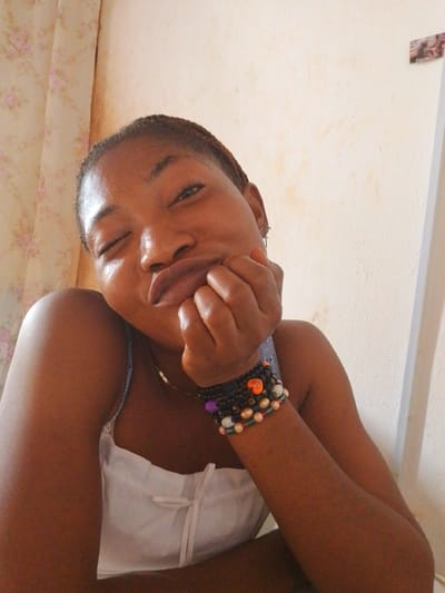 A young dark-skinned woman is in an indoor setting, possibly a bedroom. She is resting her head on her hand, wearing bracelets on her left wrist, and has on a white top. There is a floral patterned curtain to the left and a plain white wall behind her. There's no discernible detail to confirm that the location is in the city of Yelwa, Nigeria.