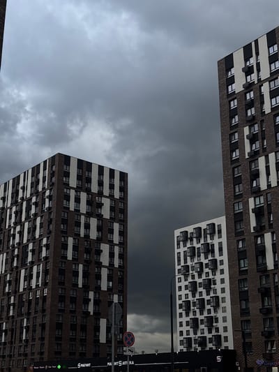 The image captures a view of residential buildings under a dark, overcast sky in Misaylovo, Russia. In the foreground, two tall, modern apartment buildings dominate the scene. The building on the left is constructed with dark brown bricks and features alternating vertical bands of white and dark gray panels. It has numerous windows, some with dark shutters. The building on the right, partially obscured by the first, is similarly designed but appears to have a slightly different pattern of white and dark vertical elements. In the middle ground, a smaller, white apartment building is visible, characterized by a facade with protruding glass-enclosed balconies arranged in a grid pattern. The sky is filled with heavy, dark gray clouds, suggesting impending rain or a storm. The lower portion of the image shows the ground level with the facades of commercial establishments, including signs for "Magnit" and "Magnit Apteka". A street lamp and traffic signs are also present. No people are visible in the image, and the scene appears to be set in an urban or suburban environment.