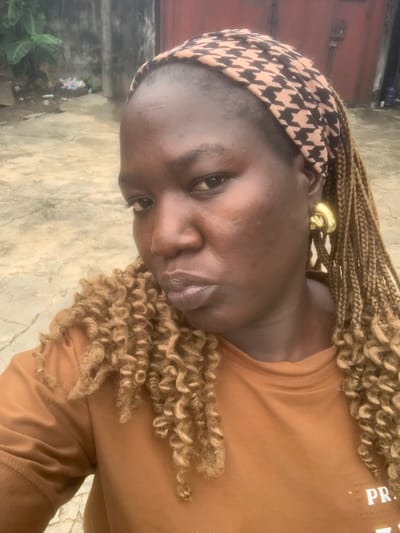 A close-up, waist-up, outdoor selfie shows a woman with dark skin, auburn braids, and large gold hoop earrings. She is wearing a brown t-shirt and a brown and black patterned headband. Her head is slightly tilted and her lips are pursed. The background shows a dirt or concrete pathway, a red wall, and some greenery. The scene appears to be in Eliozu, Nigeria.