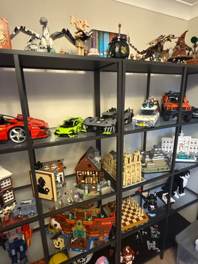 The image shows a shelf filled with various Lego constructions, arranged on black metal shelving units. The collection features a range of themes, including vehicles, buildings, and characters from popular franchises.
On the top shelf, there are Lego figures and structures that appear to be from the Harry Potter universe, including a depiction of Dobby, a cauldron, and a dragon. There are also various books visible.
The second shelf displays several Lego cars, including a red Ferrari, a lime green sports car, and a gray car. A white vehicle resembling the Ecto-1 from Ghostbusters is also present, alongside an orange off-road vehicle.
The third shelf is populated with Lego building models. One is a brown, timber-framed house. Another is a yellow, ornate building that resembles Notre Dame Cathedral. A Japanese-style castle structure is also visible. A black and white cat figure is positioned on the right side of this shelf.
The fourth shelf contains a Lego chess set with gold and brown pieces. A black dragon figure, possibly Toothless from How to Train Your Dragon, is positioned near the chess set. Further to the right, there are more Lego constructions, including a black and white cat, and a smaller white vehicle.
The bottom shelf has additional Lego creations. A large red and yellow pirate ship dominates the center. To the left, a blue and red Transformer robot is visible, along with a gray van. There are also smaller Lego building sets and figures scattered across this shelf. A yellow bucket and a skull-shaped object are also present.
The overall environment is an indoor setting, likely a room with a light-colored wall and wooden flooring partially visible at the bottom. The lighting suggests an indoor environment, possibly a home or a retail display. The specific location of Newcastle-under-Lyme, United Kingdom, cannot be determined from the visual information provided in the image.