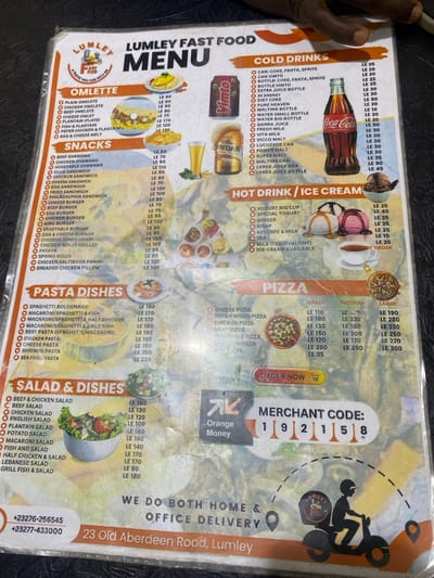 A close-up image displays a laminated menu from "Lumley Fast Food," held by a visible hand on the left. The menu is divided into several sections, including "Omelette," "Snacks," "Cold Drinks," "Hot Drink / Ice Cream," "Pizza," "Pasta Dishes," and "Salad & Dishes." Each section lists various food and beverage items with corresponding prices denominated in "LE" (Sierra Leonean Leone).

Notable text details include the "Lumley Fast Food" logo with the tagline "YOU CAN BELIEVE OUR TASTE." The menu also features contact phone numbers: "+23276-256545" and "+23277-433000," along with the address "23 Old Aberdeen Road, Lumley." The restaurant advertises "WE DO BOTH HOME & OFFICE DELIVERY." A payment option for "Orange Money" is indicated, complete with a "MERCHANT CODE: 192158." Small illustrative photos of various dishes, such as pizzas, omelettes, and salads, are integrated into the menu's design. The menu itself shows signs of use, with some reflections and minor wear. The background suggests it is placed on a dark, possibly textured surface. The overall context points to a fast-food establishment in Freetown, Sierra Leone.