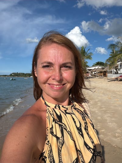 The media file shows a close-up, selfie-style shot of a woman on a beach. She is smiling and looking towards the camera. Her hair is brown and reaches her shoulders. She is wearing a yellow and black patterned top.
The background features a sandy beach with palm trees and buildings that appear to be beach bungalows. The ocean is visible in the lower part of the frame, with gentle waves lapping at the shore. Several small boats are scattered on the water. The sky is blue with scattered white clouds, suggesting a sunny day. The overall impression is one of a relaxed vacation or holiday in a tropical location. The time of day appears to be daytime, likely afternoon given the lighting. The location context provided is Baan Mae Nam, Thailand, which aligns with the visual cues of a tropical beach setting. No visible text is present in the image.