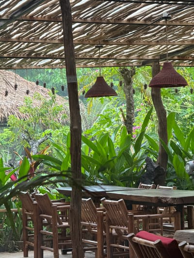 An outdoor seating area with wooden tables and chairs is set beneath a thatched roof supported by wooden posts. The area is surrounded by lush green tropical foliage, with a thatched-roof structure visible in the background. Two conical lamps hang from the ceiling, and pine cones are strung along cords as decoration. The scene appears to be in Nam Hoa Lư Ward, Vietnam, given the architectural style and vegetation.