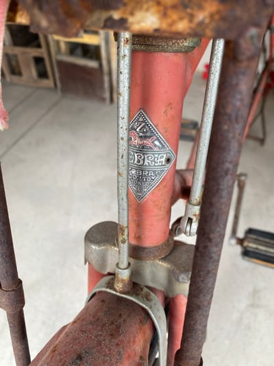 The image is a close-up, eye-level shot of a vintage red bicycle frame, focusing on a diamond-shaped metal emblem attached to the head tube. The emblem features a stylized zebra illustration at the top and lettering that appears to read "ZEBRA CO., LTD. TOKYO". The bicycle exhibits significant rust and wear on its metal components, including the frame, handlebars, and spokes. A thin pink fabric is visible on the upper left edge of the frame. The background is out of focus, showing what appears to be an outdoor setting with a light-colored, textured surface, possibly a driveway or patio, and hints of wooden structures. The scene is likely located in Baan Hua Thanon, Thailand, due to the provided context.