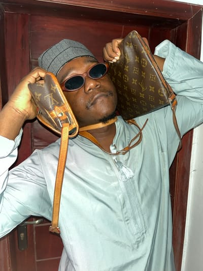 An adult male is depicted indoors in the city of Joma, Ghana. The subject, with dark skin, a goatee, and mustache, is shown from the chest up, looking upward. He wears a grey kufi, oval dark sunglasses, and a light blue-green garment with chest embroidery and a tassel. Two Louis Vuitton monogram-patterned bags are held by his hands, one positioned near each ear. The light brown leather straps of both bags are wrapped around his neck. The background consists of a dark brown wooden door with a silver handle and keyhole on the left, indicating an interior setting.