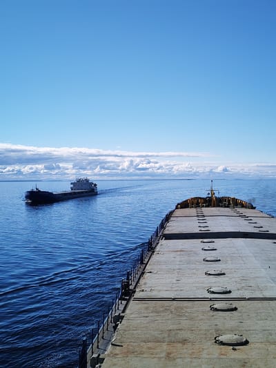 The image shows a wide-angle view from the deck of a barge, looking forward. The barge's deck is gray and textured, featuring numerous evenly spaced manhole covers. A smaller cargo ship is visible in the distance, moving in the same direction as the barge. The water is deep blue with visible waves and wakes from the vessels. The sky is mostly clear blue with some white, puffy clouds on the horizon. The location of the scene is likely on a large body of water, given the presence of the ships and horizon line. There is no visible indication of the location being in the city of Novaya Svobodka, Russia.