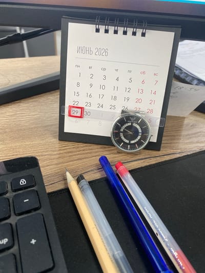 The image displays a desk with various items, including a desk calendar for June 2026, an Emporio Armani watch, a keyboard, a pencil, and three pens (one blue, one clear with a red tip, and one clear with a blue tip). The calendar is open to June 2026, and the 29th is highlighted with a red marker. The watch is resting on the calendar. The keyboard is partially visible on the left side of the frame. The background includes a monitor and a wooden desk surface. The lighting suggests an indoor setting, likely an office or home workspace. No people are visible in the image. There is no discernible information in the image that would indicate a specific location in Kaspiysk, Russia.