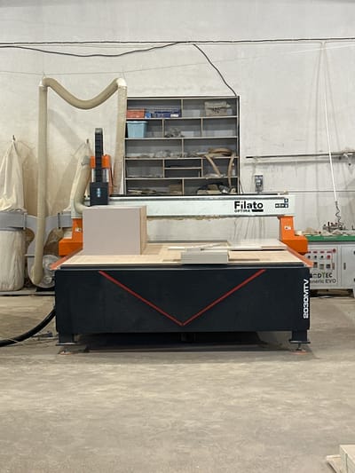The image shows a CNC router machine in a workshop. The machine is predominantly black and orange, with a long, white gantry labeled "Filato OPTIMA." A control panel with various buttons and indicators is visible on the top right of the machine. The machine is positioned against a concrete wall, and a large roll of fabric or protective covering is seen at the bottom of the image. There are also several flexible ducts connected to the machine, likely for dust collection.

The overall scene appears to be a manufacturing or fabrication setting. The lighting suggests it's indoors, and there are no obvious cues for time of day or weather. The location is identified as Kizilyurt, Russia. There is no visible text on the main subject of the image, apart from the labels on the machine itself. The image is taken from a side angle, with the machine taking up most of the frame. There are no people visible in the image, nor any specific activity or event taking place. The focus is solely on the industrial machinery.