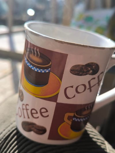 A white ceramic mug with a handle is depicted in a close-up, angled shot in Dolistovo, Bulgaria. The mug features a decorative pattern of brown and white checkered squares. Within these squares, illustrations include stylized brown coffee cups with blue and white checkered bands on yellow/orange saucers, individual brown coffee beans, and the word "Coffee" in brown text. Steam is visibly rising from the interior of the mug, indicating a hot beverage. The mug's rim shows minor chips and wear. It rests on a dark, ribbed textile surface, possibly clothing. The background is heavily blurred with bright light sources and indistinct shapes, suggesting an outdoor or semi-outdoor environment under direct sunlight. No individuals are visible.