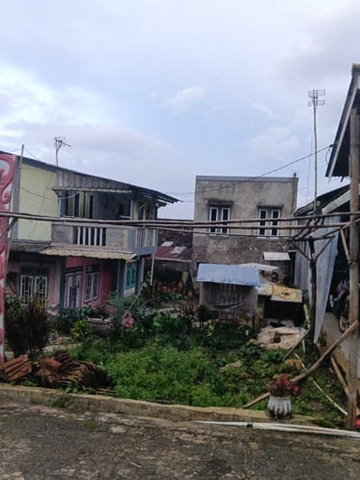 This media file captures a residential scene in what appears to be a densely settled area, consistent with An Khe Ward, Vietnam. The image is taken during the daytime, likely under an overcast sky, as indicated by the diffused light and lack of harsh shadows. No people are visible, and no specific activity is taking place.
The foreground features a weathered asphalt or concrete path, beyond which is a strip of overgrown green grass and various plants. To the left, there's a pile of brown, interlocking roof tiles. In the mid-ground, multiple buildings are visible. On the far left, a two-story house with a light yellow upper section and a pink lower section stands out. It has a balcony with white railings on the second floor and pink-framed doors or windows on the first floor, along with a prominent red, decorative element on its left side.
Behind and to the right of this house, another two-story building with a plain, weathered concrete or stucco facade is visible. It features two windows on the upper floor with white frames. Utility wires stretch across the scene, connecting to various structures. Two distinct television antennas are perched on rooftops โ one on the left building and another on the far right.
Between the buildings and in the mid-ground, there's a significant amount of lush green vegetation and what appears to be a small, makeshift shelter with a blue tarp roof. On the far right, a structure made of bamboo poles creates a partial frame, with a motorcycle dimly visible behind it. A white, rustic pot with red flowers sits on the ground in the lower right. The overall impression is one of an established but possibly developing neighborhood with a mix of permanent and improvised structures. No visible text can be discerned in the image.
