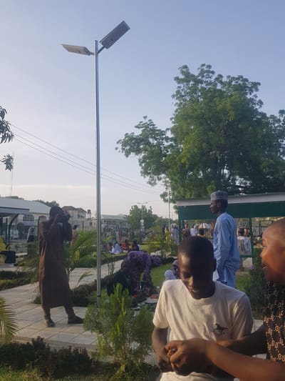 A medium, eye-level outdoor shot shows Muslim men in Bauchi, Nigeria, kneeling and praying on outdoor prayer mats arranged over patterned white concrete. The sun shines brightly. A few of the men wear Muslim prayer caps or traditional Muslim clothing, one man in a blue, long-sleeved shirt and another in brown robes. In the foreground are two men, one in a white t-shirt with a horse design and the other in a black, patterned shirt. A tree and a solar lamp post are prominent features in the background. In the far background are buildings and a tree line. Overhead electric lines extend towards the foreground.