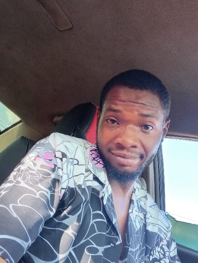 A man with short black hair and a dark beard wears a patterned shirt with black, gray, and pink floral designs. He has brown eyes and is looking directly at the camera. The background suggests he is inside a vehicle, with brown fabric upholstery visible behind him and a glimpse of sky through a window. The image is rotated, so the ceiling appears to be on the right side.