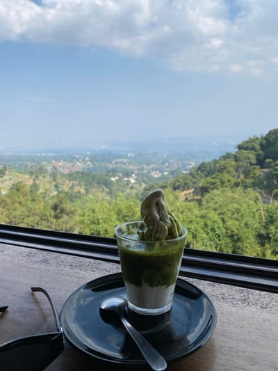 A matcha dessert is presented in a clear glass on a black plate. The dessert has layers of white and green, topped with a swirl of green matcha soft serve. A spoon rests on the plate next to the glass. The setting appears to be a restaurant or cafe with a large window offering a view of a lush, green landscape with hills and a distant city. The sky is blue with scattered white clouds. The table the dessert is on is made of dark wood. The image is taken from a slightly elevated angle, looking towards the view. The general location is Kayuambon, Indonesia.