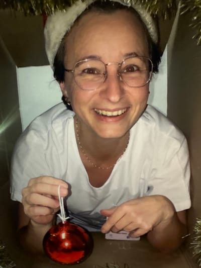 A light-skinned adult female is depicted from the chest up, positioned inside a brown corrugated cardboard box. She wears a white short-sleeved t-shirt, a delicate light-colored beaded necklace, and a white and red Santa hat. Her short, dark hair is partially visible beneath the hat. She has brown eyes and wears round, wire-frame glasses. Her mouth is open in a wide smile, revealing teeth. Her left hand holds a large, shiny red spherical Christmas ornament by its white string hanger. Her right hand rests inside the box near a small, light-colored object, possibly a label or tag, and metallic strands. Green and gold tinsel decorates the upper edge of the cardboard box, partially framing her head. The lighting appears soft, creating reflections on the ornament and glasses. The scene is located in Tourcoing, France.