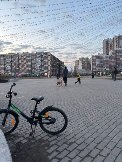 This image depicts an urban square in Kudrovo, Russia, under an overcast sky with strung lights overhead, likely at dusk or dawn.

In the foreground, a black "DENTON" brand bicycle with orange wheel reflectors and an extended kickstand is parked on a grey paved surface, partially obscured by a low concrete structure on the left. The paving consists of rectangular grey pavers arranged in a basketweave pattern, with damp patches visible near the bicycle.

In the midground, a person wearing a dark jacket and trousers walks from right to left, leading a brown and white Beagle dog on a leash. To their right, a child in a bright yellow jacket and dark trousers runs towards the right. Further right, another individual in a dark jacket and blue jeans stands, facing left. Several other indistinct figures are visible in the background, walking or standing across the square.

The background features multiple large, modern residential buildings with numerous windows and balconies. The building on the far left exhibits a facade with a patterned mix of brown, orange, grey, and white panels. Buildings on the right have dark window frames and red-brown accent panels. Overhead, numerous strings of small lights are suspended horizontally, spanning the entire visible sky and creating a grid-like pattern against the pale, partially cloudy sky. The horizon shows a subtle orange tint.