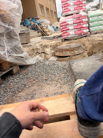 The image depicts a construction site in Příbram, Czechia. In the foreground, a person's hand is visible holding a small object, possibly a cigarette butt. The person is wearing blue work pants and a dirty work boot. The environment is rough, with gravel, dirt, and construction materials scattered around. In the background, stacks of cement bags and building materials are present, along with a section of a building. A metal manhole cover is also visible, partially submerged in the dirt. The scene suggests ongoing construction or renovation work.