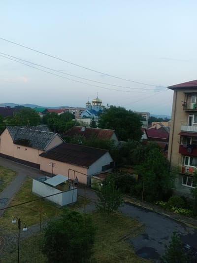 The image depicts a view of an urban or suburban setting in Tiachiv, Ukraine, likely during the late afternoon or early evening due to the soft, diffused light and overcast sky. Several residential buildings of varying sizes and architectural styles are visible, including a multi-story building with balconies on the right, and smaller, single-story houses with pitched roofs. In the background, a prominent church with golden domes stands out against the skyline, indicating a religious presence in the area.
The scene is characterized by a mix of developed structures and natural elements. Trees and green spaces are interspersed among the buildings, suggesting a somewhat verdant environment. Utility lines are strung across the sky, typical of a populated area. In the foreground, there is a paved pathway and a patch of overgrown grass. A small, utilitarian structure with a corrugated metal roof is situated on the left, and two individuals can be seen near it. One person appears to be standing and observing, while another is closer to the structure. A lamppost with two lights is visible on the left side of the frame, further contributing to the urban setting. The overall mood is peaceful and everyday, with no indication of any specific event or dramatic activity. The weather appears to be calm, with no strong visual cues of wind or rain.