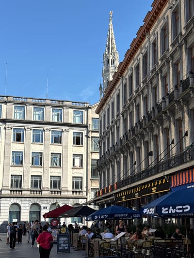 This is a wide shot of the Grand Place in Brussels, Belgium, on a bright, clear day. The Grand Place is a historic market square and a UNESCO World Heritage site. In the background, the iconic spire of the Brussels Town Hall dominates the skyline. The foreground is filled with people, tables, and umbrellas from outdoor cafes. The buildings surrounding the square exhibit intricate architecture with ornate facades, balconies, and numerous windows. People are walking, dining, and socializing in the bustling square. Several restaurants are visible with outdoor seating, indicated by tables, chairs, and umbrellas. The atmosphere is lively and inviting, capturing the essence of a European city square.