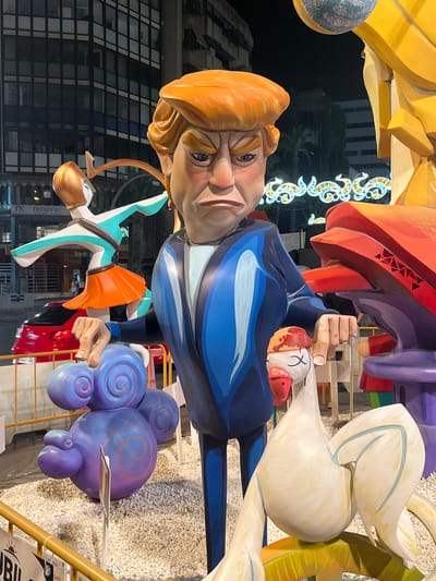 The image shows a street scene at night in Alicante, Spain, featuring a large, colorful satirical sculpture. The sculpture depicts a caricatured figure resembling Donald Trump, wearing a blue suit with light blue accents and sporting his signature orange hair. His expression is stern and his brow is furrowed. He is positioned centrally, with his hands extended outwards.
To the left of the Trump figure, a stylized, light-skinned dancer in a turquoise and white outfit with an orange skirt is depicted in a dynamic pose. Further left, a red car is visible, with its headlights on, parked on a street. Behind the sculpture and car, a modern building with a grid of dark windows is illuminated.
In front of the Trump figure, to his left, is a large, multi-lobed purple sculpture with swirling patterns. To his right, a large white bird figure with its eyes crossed and a red beak is depicted. This bird appears to be part of the larger sculpture. In the background, festive lights are strung across the street. The ground is covered in small white pebbles. Signs are visible in the foreground, with partial text suggesting a public event.