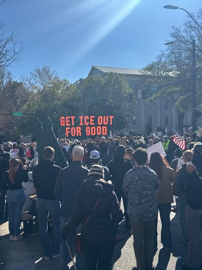 A daytime street protest features a dense crowd. A large electronic sign displays the message "GET ICE OUT FOR GOOD." The US flag is visible within the crowd. People hold signs and march down the street. The architecture of a tall building can be seen in the background with bare trees. "E Broad St" is written on the green sign on the street.