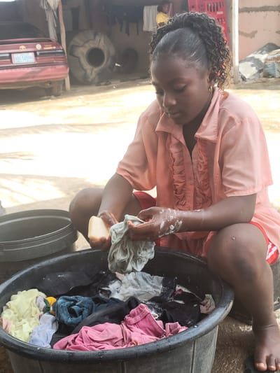 A woman washes clothes in a black plastic tub in Madalla, Nigeria. The woman, wearing a pink blouse and red shorts, kneels beside the tub, scrubbing a grey garment with a bar of soap. The tub contains various clothing items in pink, black, blue, and yellow. Behind the woman, there's a building, a red car, and a large cylindrical object.